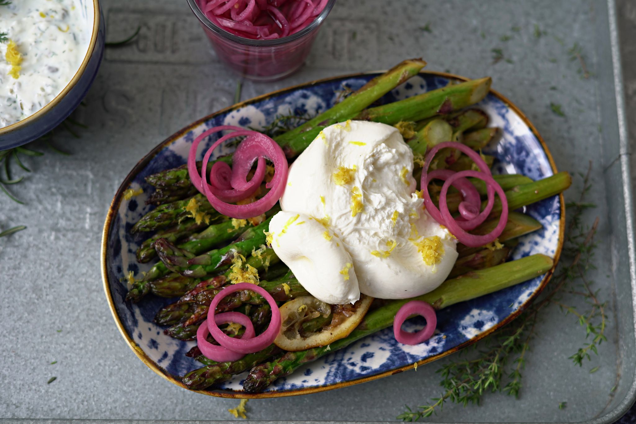 Zum Anrichten wird der Burrata über den Spargelstangen etwas auseinander gezupft und mit dem Ranchdip beträufelt. Zuvor eingelegte rote Zwiebelringe bringen Farbe auf den Teller.